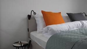 Una cama con ropa de cama blanca, una almohada naranja, una almohada gris y una colcha verde. Hay una mesita con un libro y una lámpara en la pared junto a la cama.
