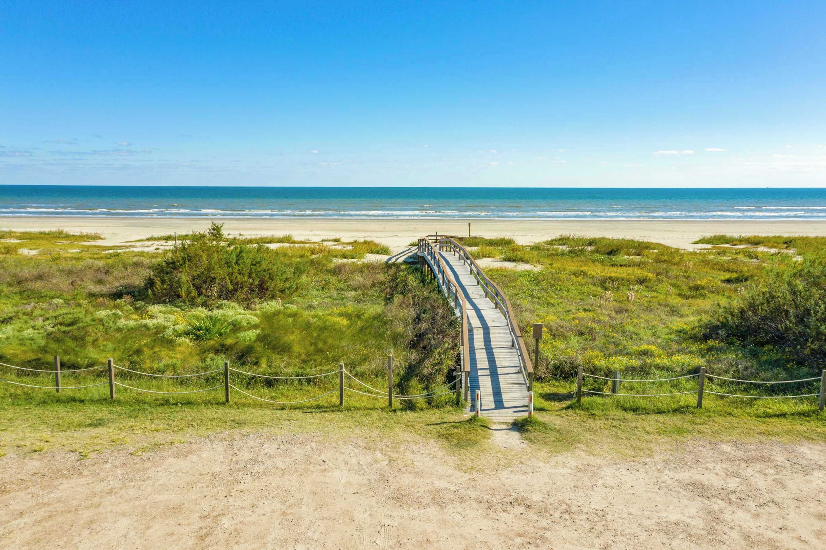 A beach with a wooden boardwalk leading to the shore, surrounded by greenery and a clear sky.
