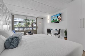 A modern hotel room with a large bed, a desk with two chairs, a television mounted on the wall, and a window with a view of palm trees. - Bright Retreat Near Disney + Free Parking | Orl (Kissimmee)