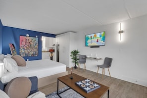 Ein modernes Hotelzimmer mit einem Bett, einem Fernseher, einem Essbereich mit Stühlen und einer Küche im Hintergrund.