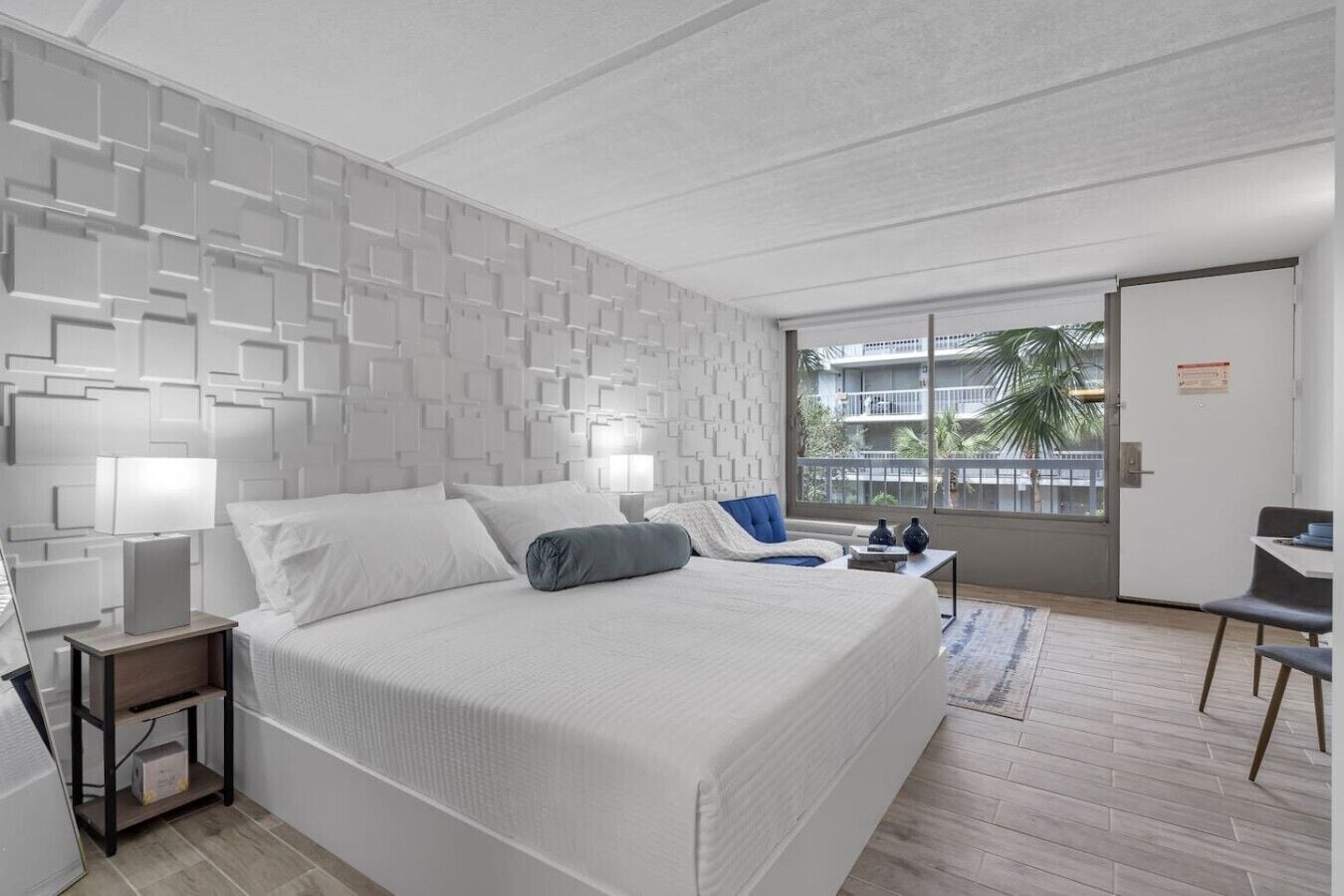 A modern hotel room with a large bed, bedside tables, a desk, and a window with a view of buildings and palm trees.