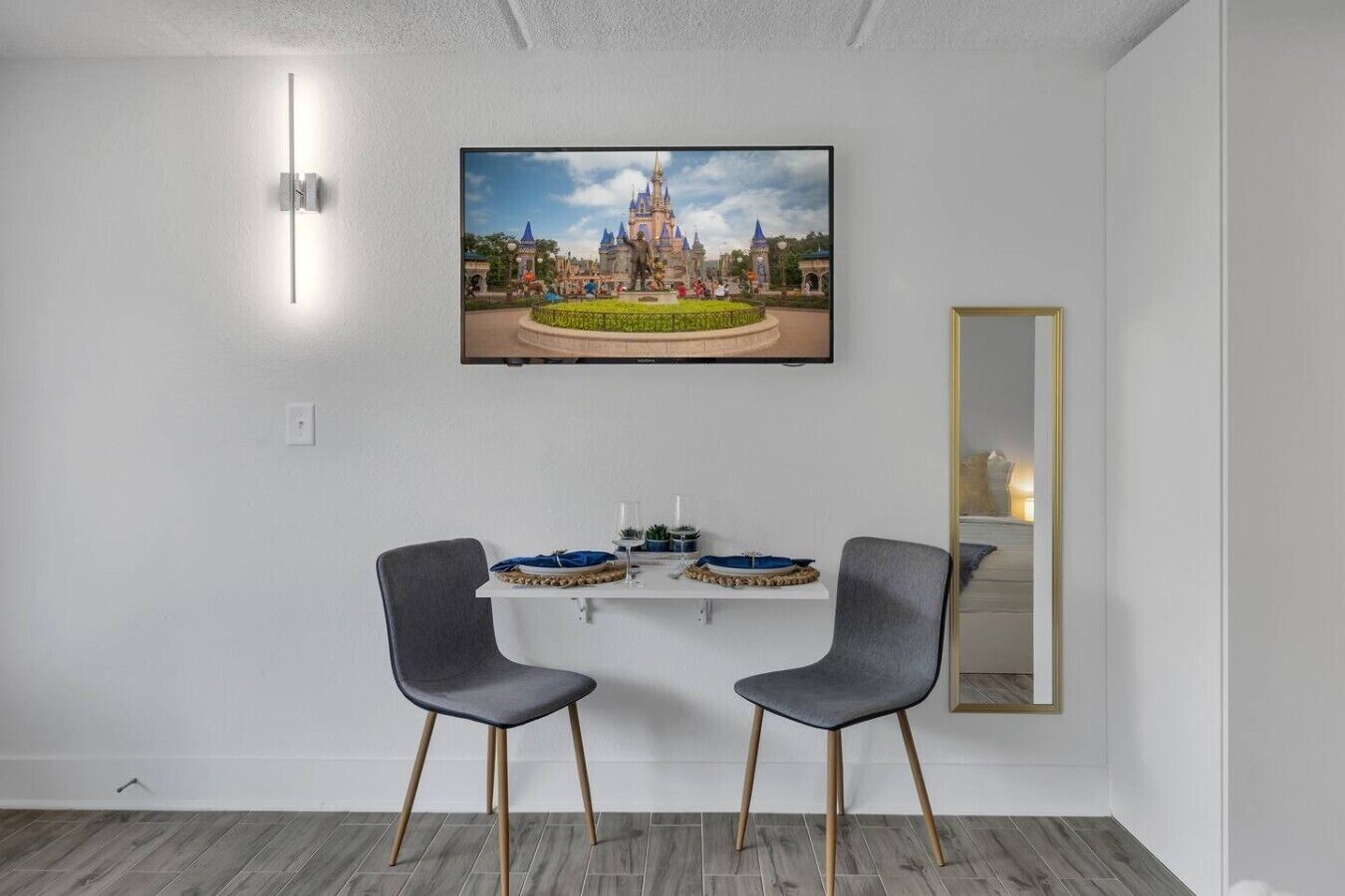 Quarto de hotel com televisão de tela plana na parede, duas cadeiras cinzas, uma mesa pequena com bandeja decorativa e espelho com moldura dourada.