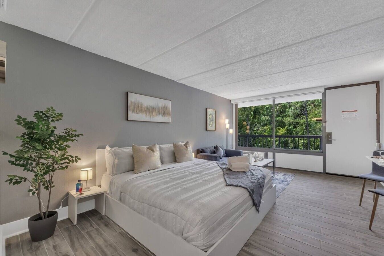A modern bedroom with a large bed, bedside tables, a potted plant, and a view of greenery through a large window.