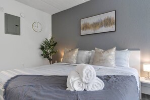 A hotel room with a bed, pillows, a clock, a plant, and a painting on the wall. - Cozy Family Escape Near Disney + Parking | Orl (Kissimmee)