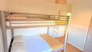 Een stapelbed met een witte matras en een geboeide deken, een hanglamp van wilgentenen en een houten vloer.