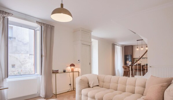 A living room with a sofa, a table with books, a lamp, and a view of a kitchen and dining area. - L'Élégante - Petfriendly - Proche gare (Laval)