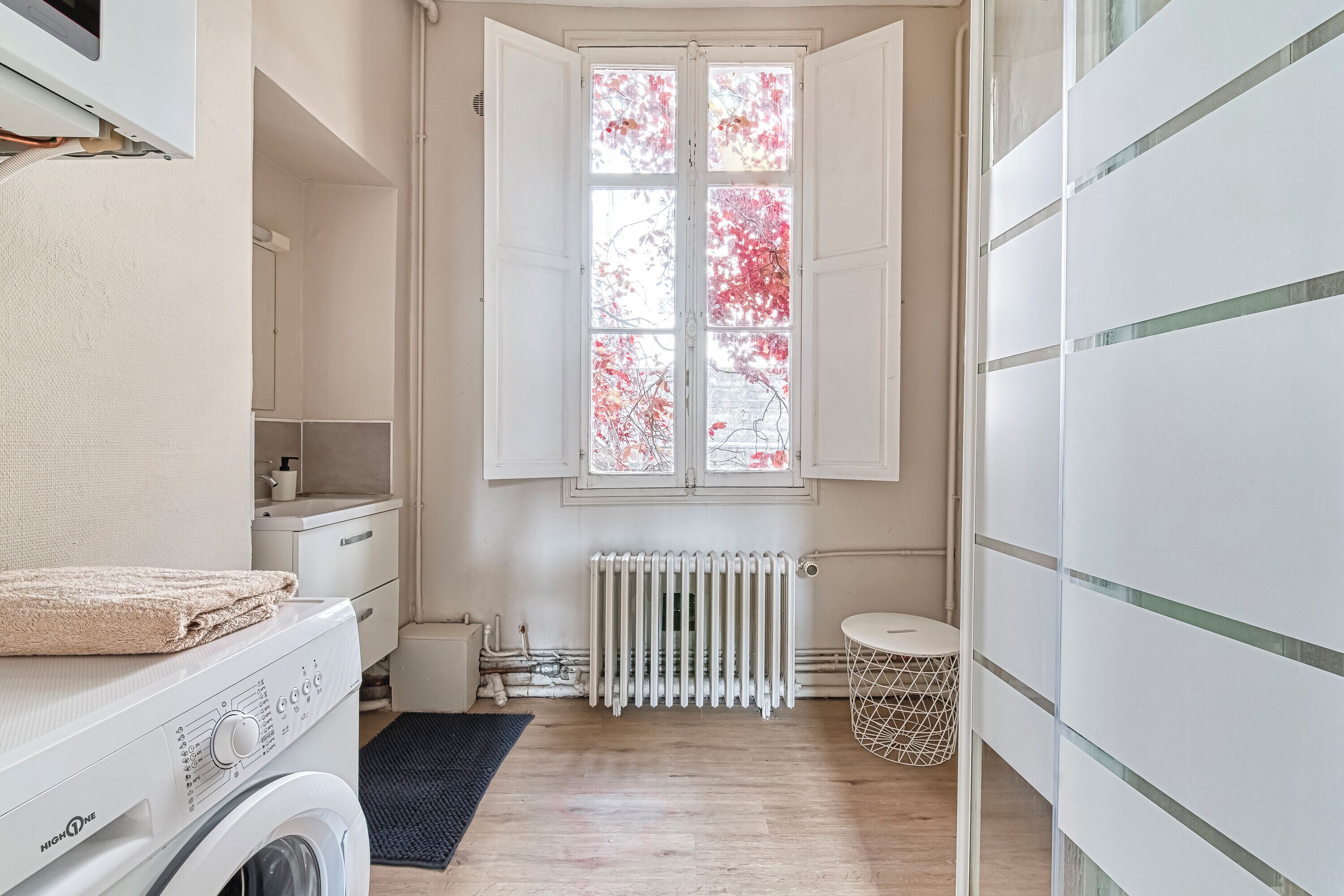 A laundry room with a washing machine, a drying rack, a radiator, and a window with a view of trees.