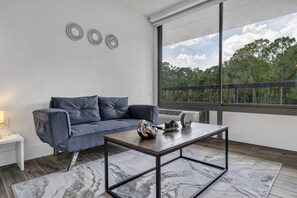 Ein modernes Wohnzimmer mit einem grauen Sofa, einem Holztisch und Blick auf Bäume durch ein großes Fenster.