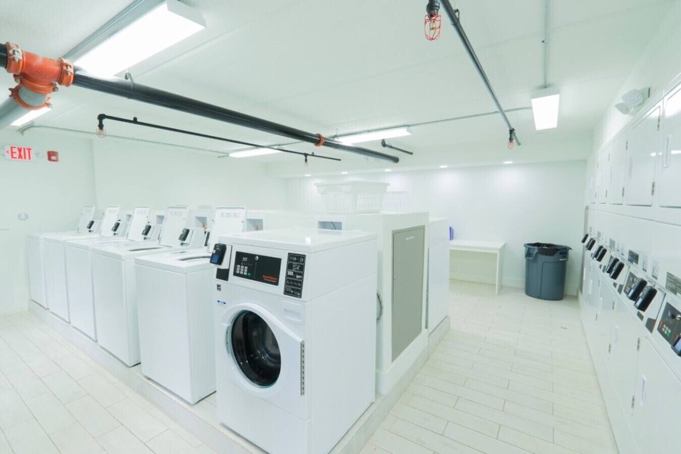 Een wasserette met meerdere wasmachines en drogers, een vluchtroute aanduiding en een afvalbak.