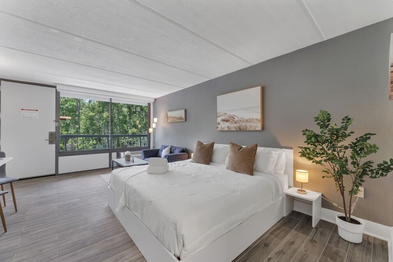 A modern bedroom with a large bed, a small sofa, a desk, and a balcony with a view of greenery.