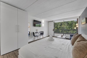 A modern hotel room with a large bed, a desk, and a balcony with a view of trees. - Resort-Style Retreat Near Disney + Parking | Orl (Kissimmee)