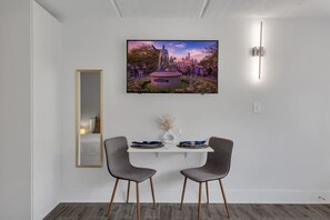 A modern dining area with a TV mounted on the wall displaying a park scene, two chairs, a table with a vase, and a mirror on the wall. - Resort-Style Retreat Near Disney + Parking | Orl (Kissimmee)