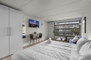 A modern hotel room with a large bed, a dining area with chairs, a TV mounted on the wall, and a balcony with a view of buildings. - Top Disney Location w Pool & Free Parking | Orl (Kissimmee)