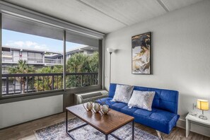 A modern living room with a blue sofa, a wooden coffee table, and a large window offering a view of a building and greenery outside. - Top Disney Location w Pool & Free Parking | Orl (Kissimmee)