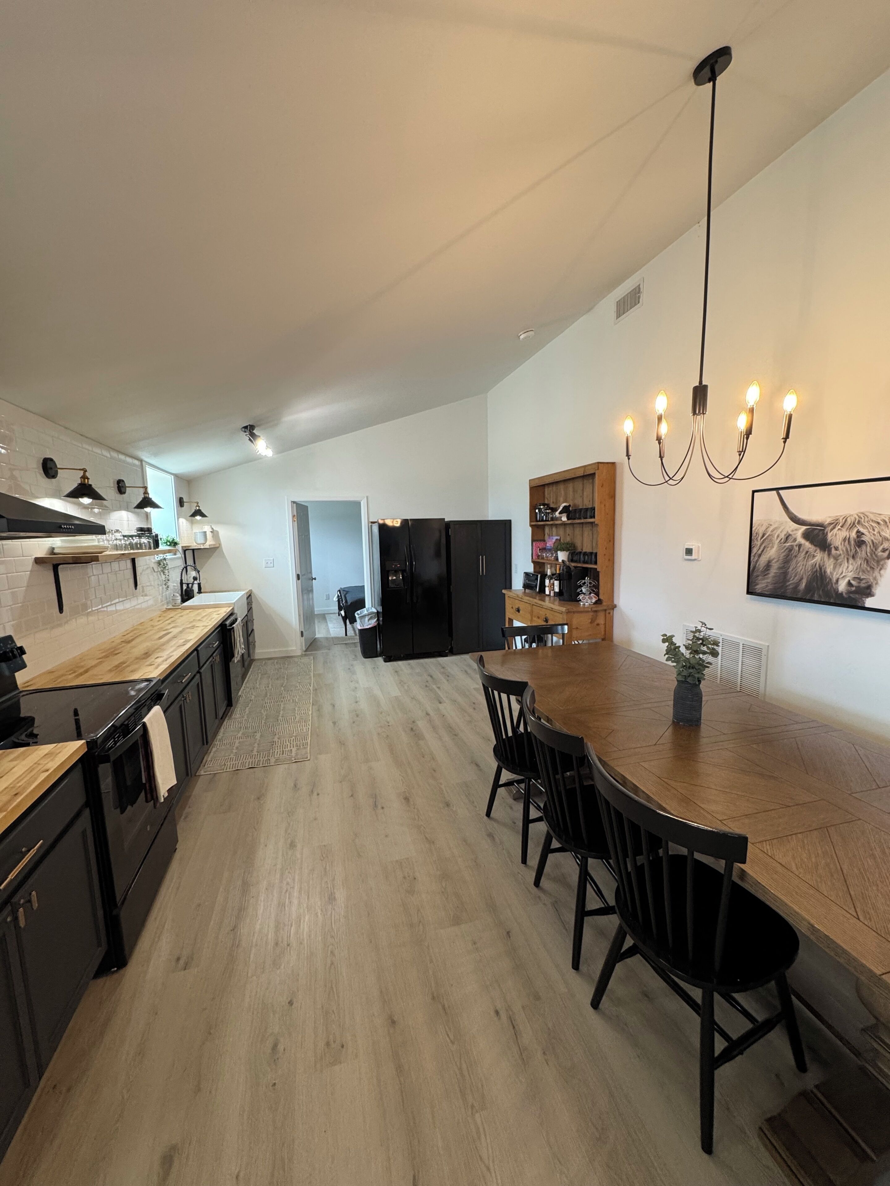 A modern kitchen with a wooden island and black chairs, a black refrigerator, and a chandelier.