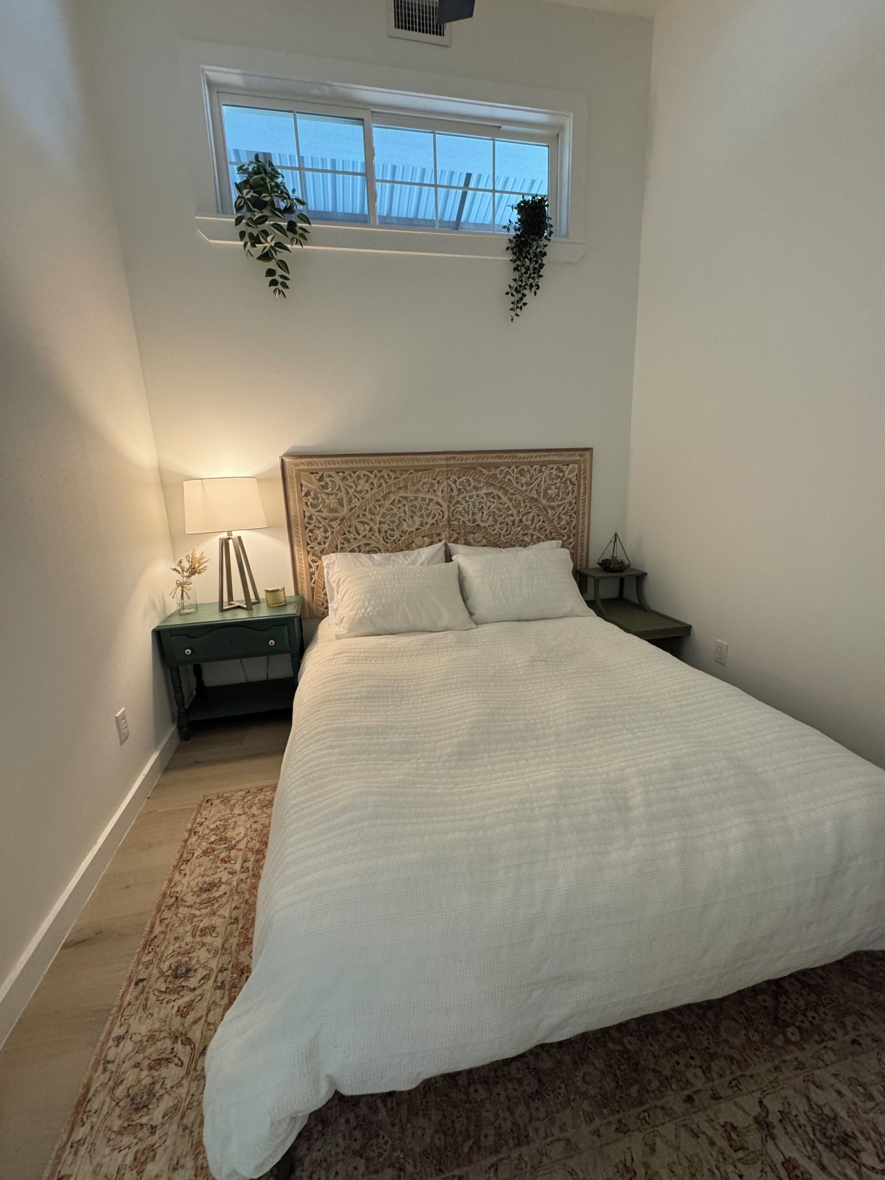 A bedroom with a large bed, a nightstand, a lamp, a window with a view, and a decorative plant.