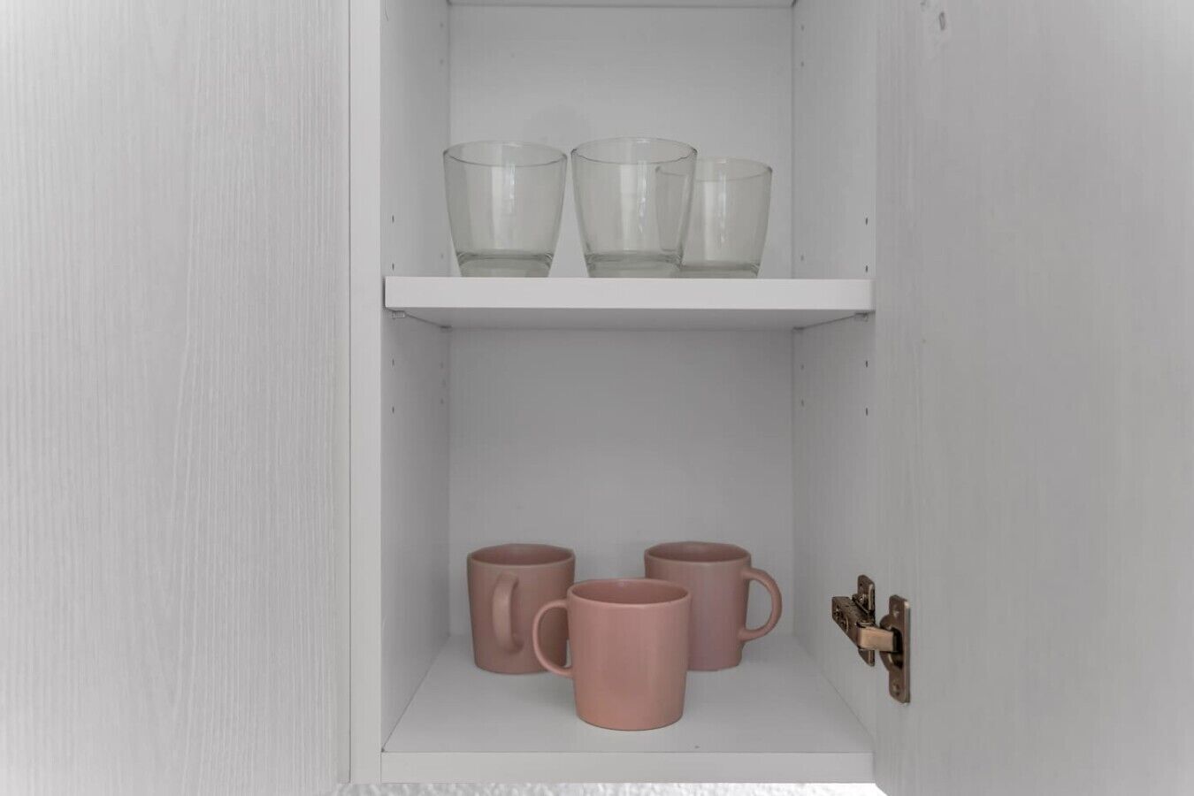 Un mueble blanco con dos estantes; el estante superior contiene tres vasos de cristal, y el estante inferior contiene tres tazas rosas.
