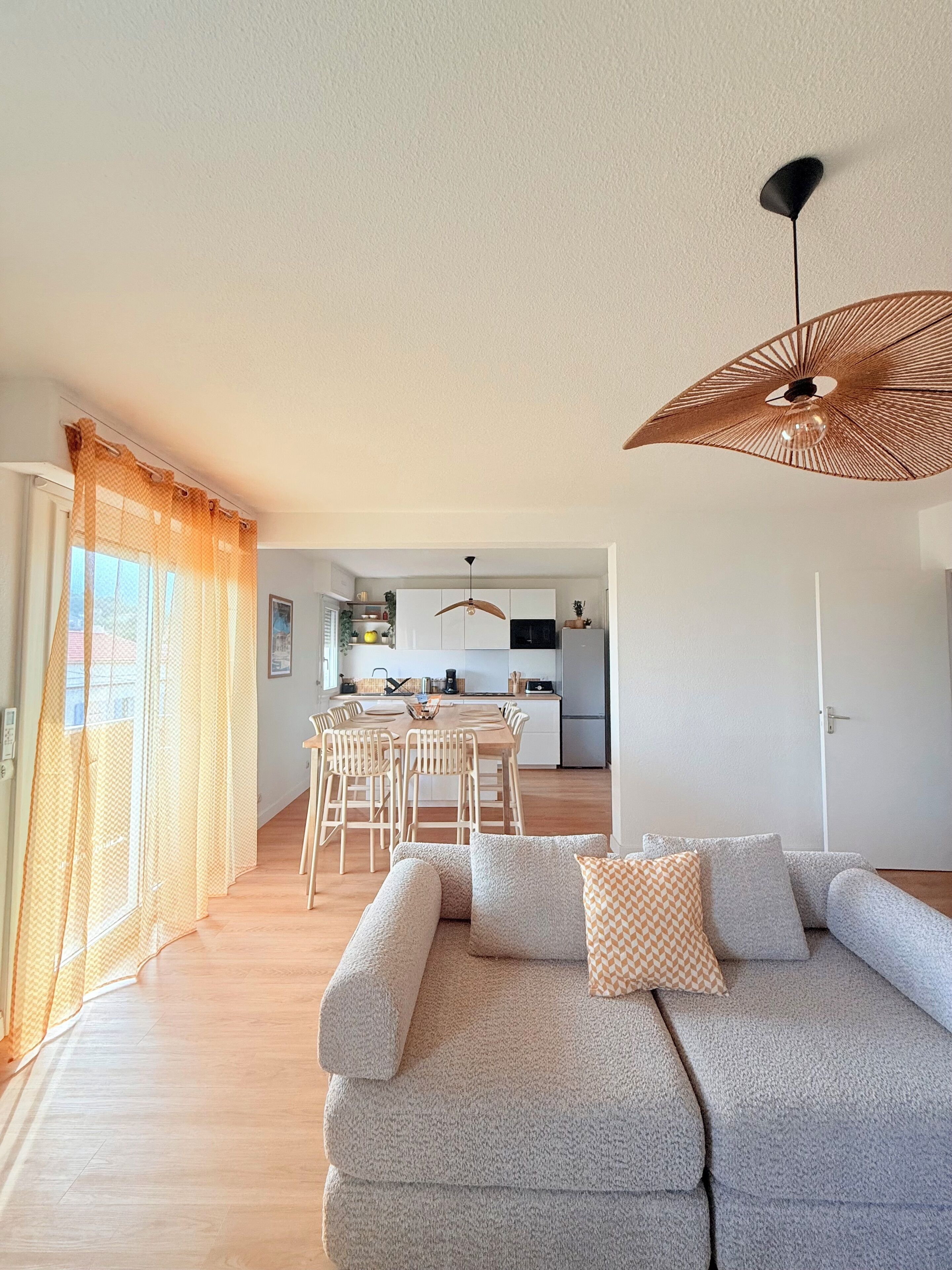 Een woonkamer met een bank, een eethoek met stoelen, een keuken op de achtergrond en een groot raam met gordijnen.