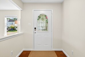 A white door with a glass panel and a doorknob, a white wall, a window with a view of a house, and a brown doormat. - Roadmap Lodging: Geist Waterfront 4BR, Fishers (Fishers)