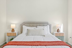 A neatly made bed with a grey upholstered headboard, white bedding, and a red throw blanket. There are two bedside tables with lamps and a small vase on each. - Roadmap Lodging: Geist Waterfront 4BR, Fishers (Fishers)