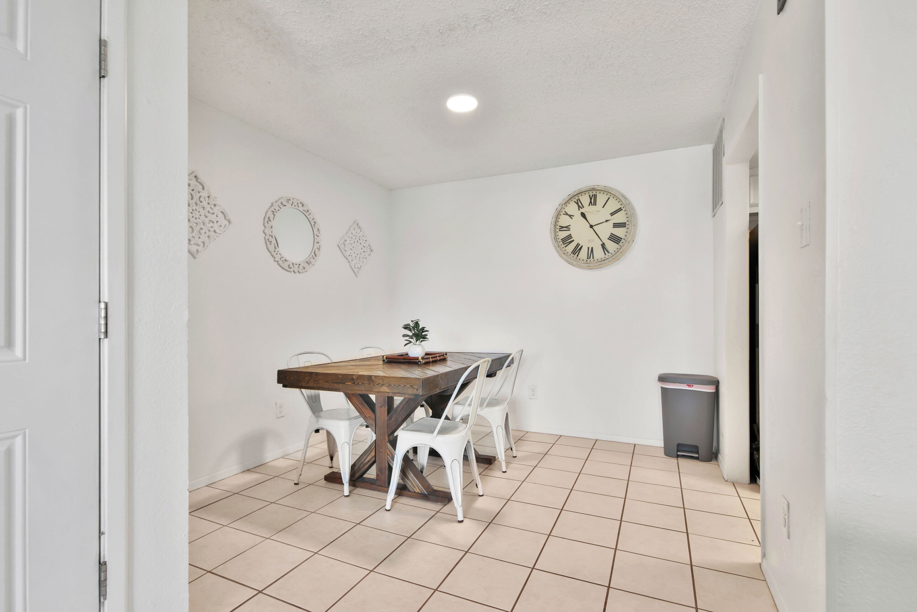 A dining area with a wooden table, white chairs, a wall clock, and a trash bin.