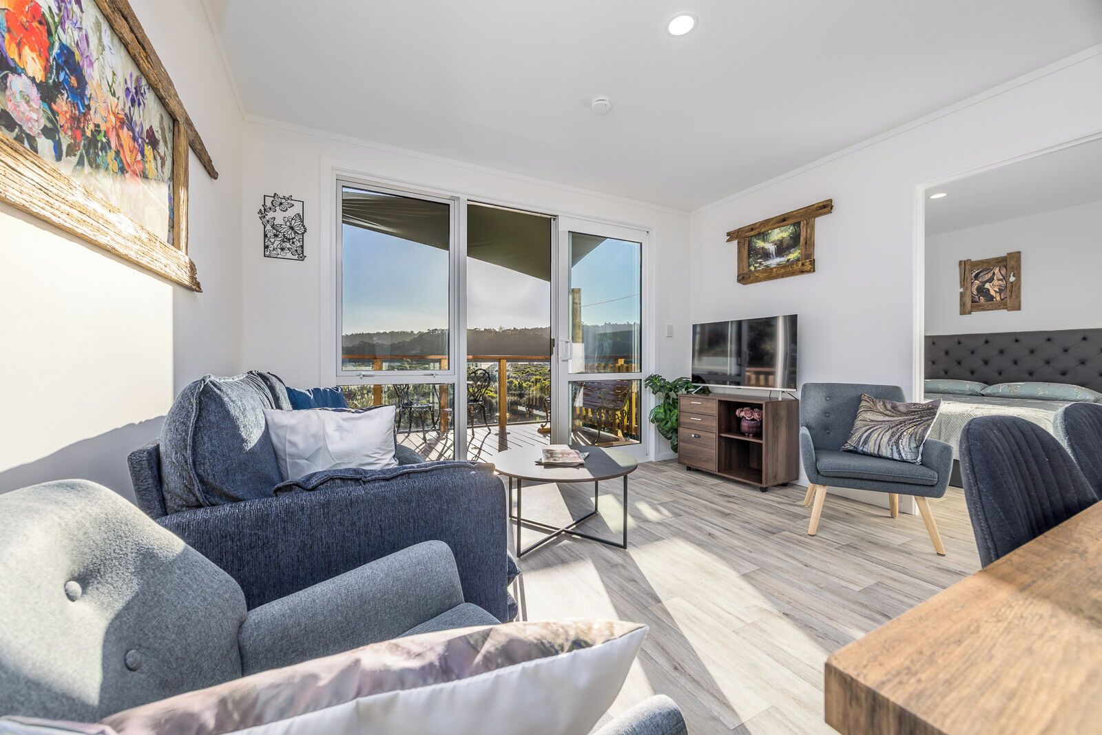 A modern living room with a sofa, a coffee table, and a TV. There is a balcony with a view of the landscape.