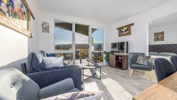 A modern living room with a sofa, a coffee table, and a TV. There is a balcony with a view of the landscape.