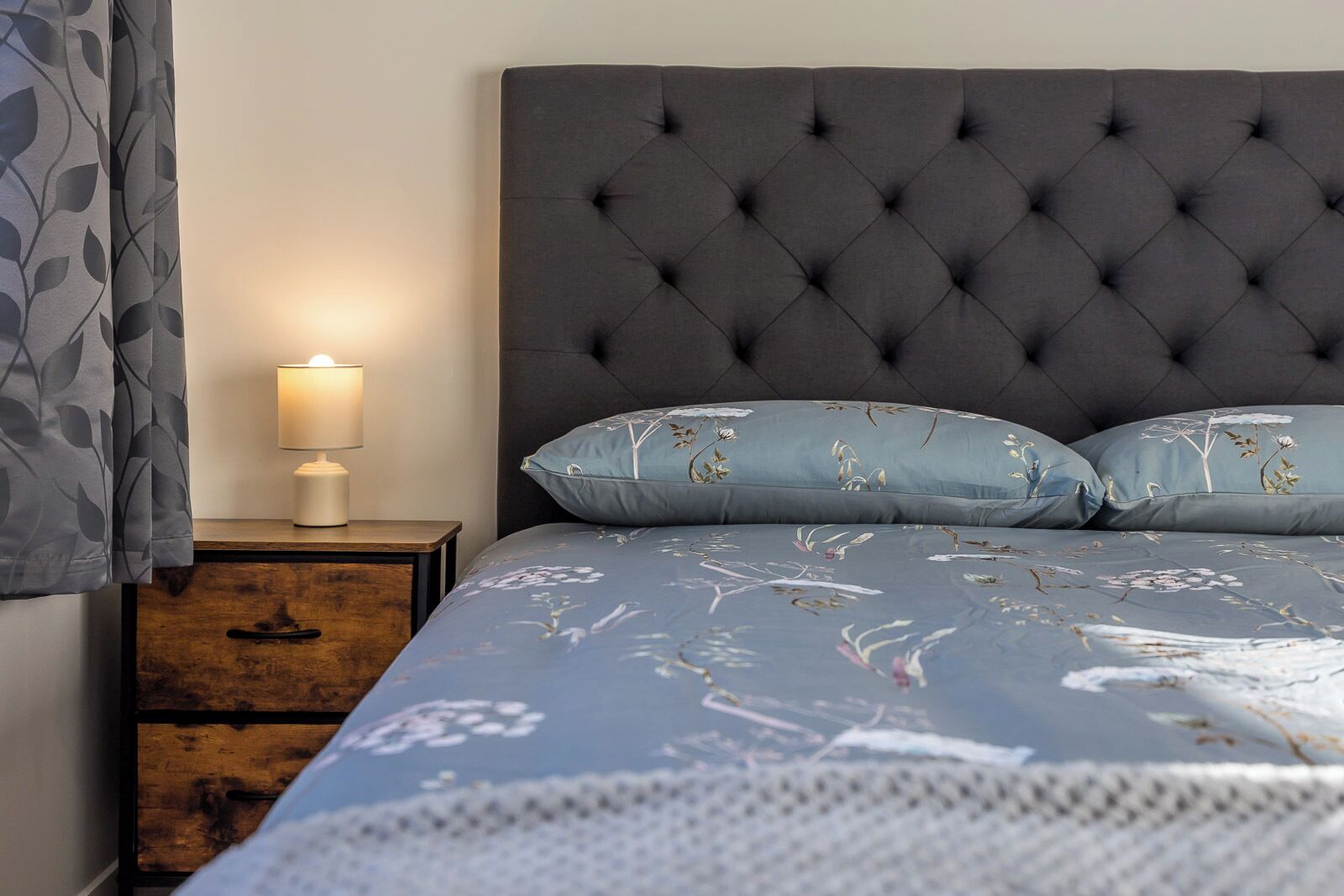 Una cama con cabecera capitonada, una mesita de noche con lámpara, y un mueble auxiliar con cajones.