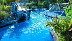 Piscina con caída de agua, rodeada de plantas tropicales y una terraza de madera.