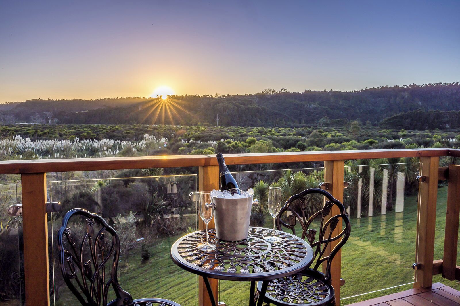 A balcony with a table and chairs, a bottle of champagne, and a glass, overlooking a landscape with trees and a sunset.