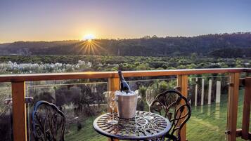 A balcony with a table and chairs, a bottle of champagne, and a glass, overlooking a landscape with trees and a sunset.