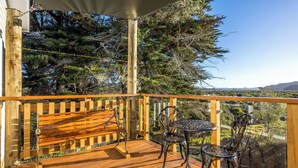 A wooden deck with a bench and metal chairs, overlooking a landscape with trees and hills.