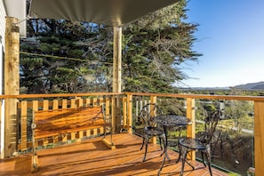 A wooden deck with a bench and metal chairs, overlooking a landscape with trees and hills.
