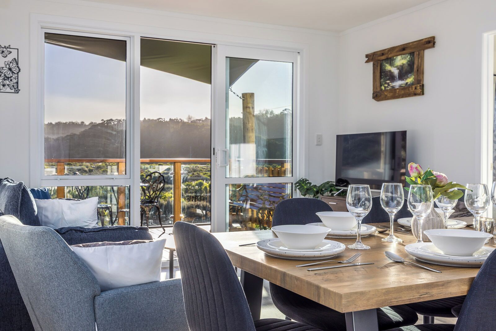 A dining area with a wooden table set for four, surrounded by chairs, with a view of the outdoors through large sliding glass doors.