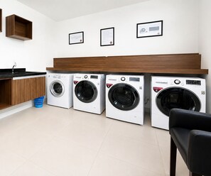 A laundry room with four front-loading washing machines, a wooden bench, and a sink. - Cozy, fully equipped studio apartment with coworking space, gym, and great location. (Curitiba)