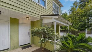A two-story house with a covered porch, a front door, and a small tree in the front yard. - Oasis on Desoto (Ocean Springs)