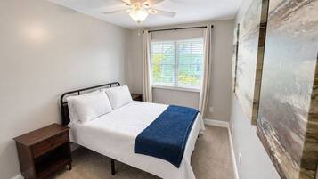 A bedroom with a bed, a nightstand, a ceiling fan, a window with blinds, and a large abstract painting on the wall.