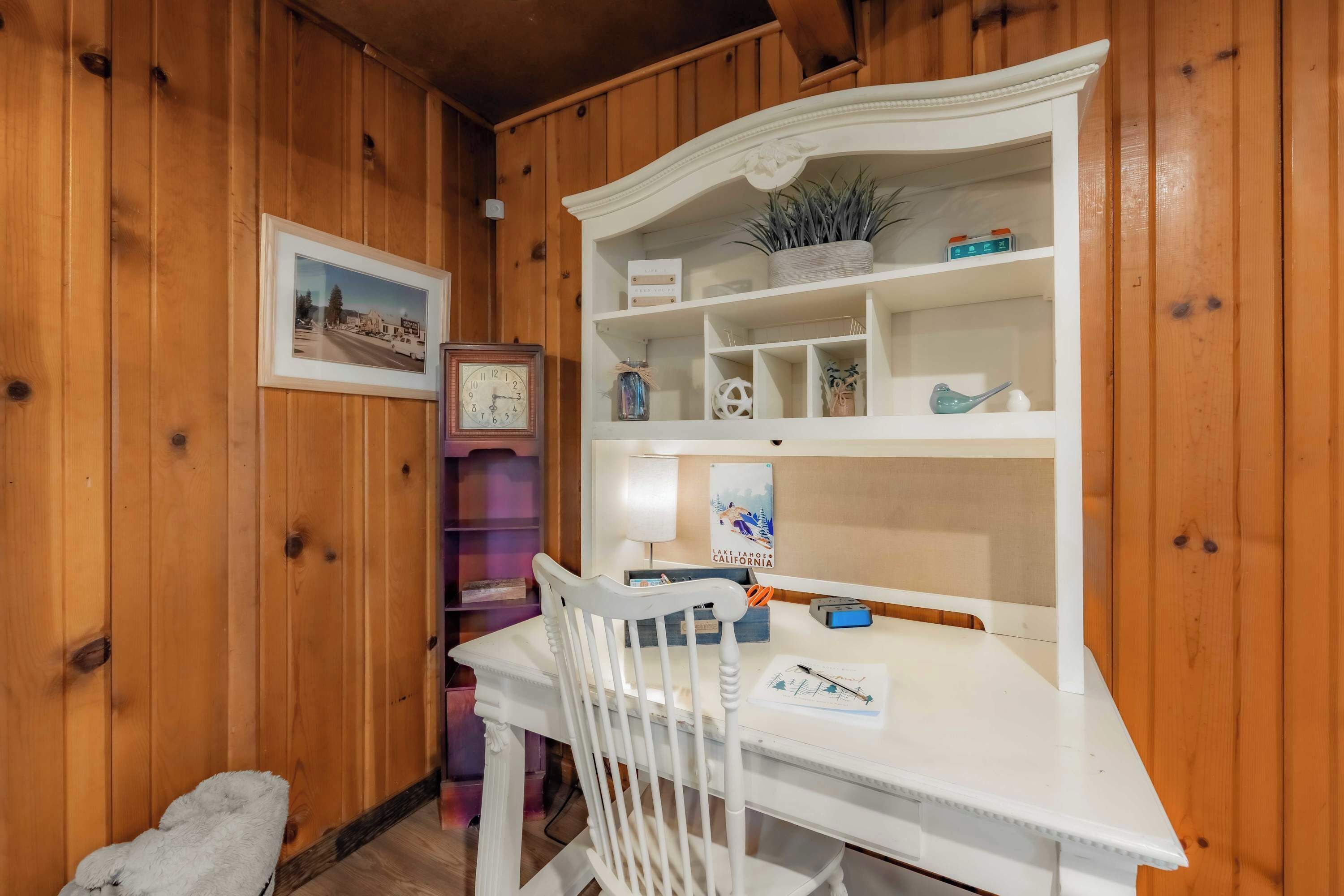 A wooden interior with a white desk, a chair, a framed picture, a clock, and a shelf with decorative items.