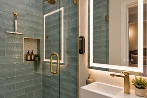 Een moderne badkamer met een douchecabine van glas, een witte wastafel met gouden details en een spiegel die een slaapkamer weerspiegelt.
