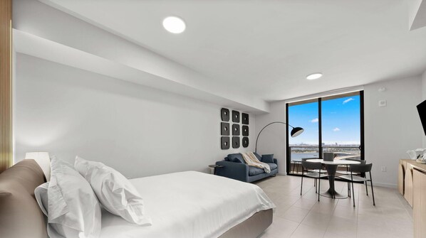 Quarto moderno com cama, sofá, mesa de jantar com cadeiras e uma janela grande com vista para a cidade.
