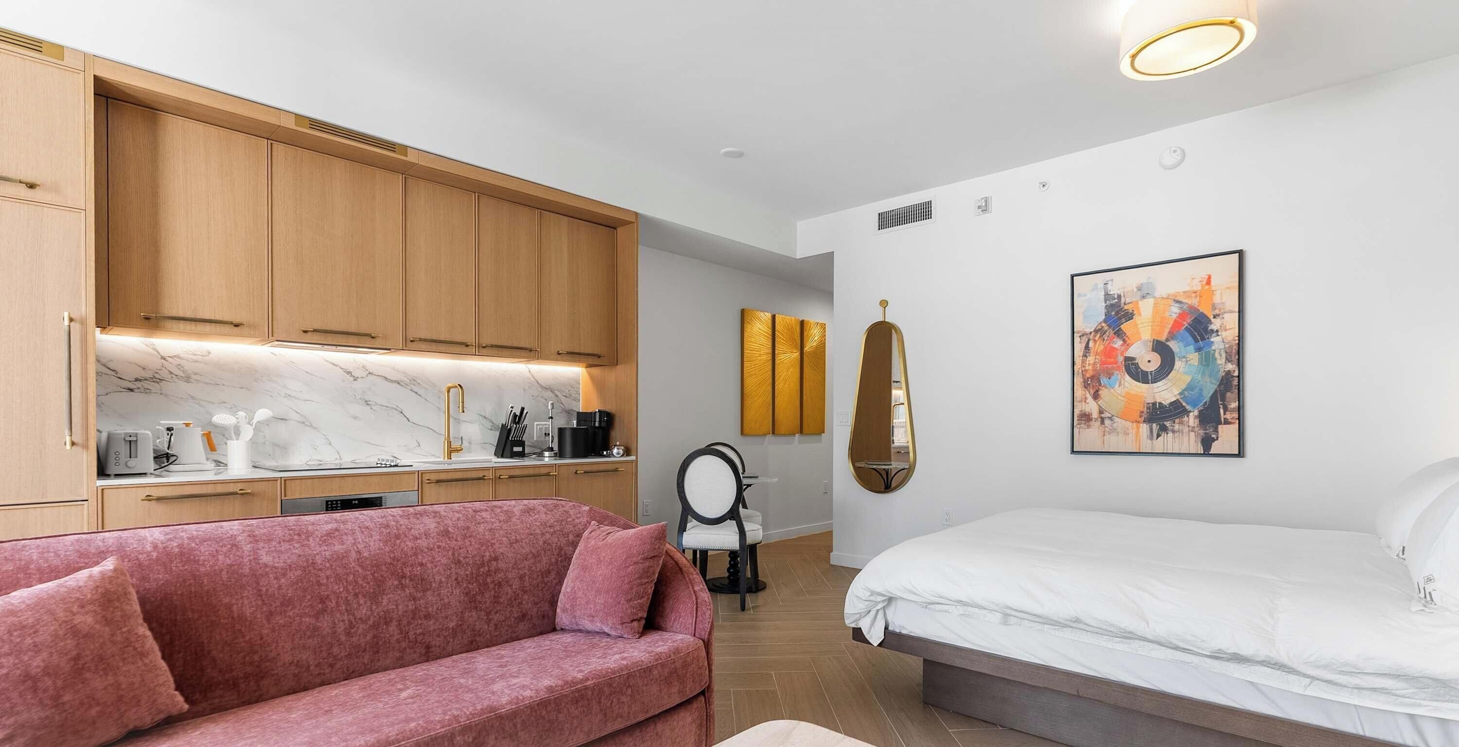 Ein modernes Schlafzimmer mit einem Bett, einem rosa Sofa, einer Küchenzeile mit Marmorarbeitsplatten und einem wandmontierten Spiegel.
