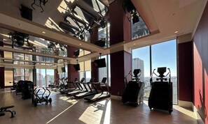 Ein Fitnessstudio mit Laufbändern, Ergometer und Krafttrainingsgeräten, großen Fenstern mit Blick auf die Stadt und einer hohen Decke mit freiliegenden Balken.