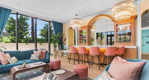 A modern living room with a sofa, coffee table, and ottomans. There is a bar area with stools and a mirror. - Rooftop Restaurant in NoMad Residences Wynwood (Miami)