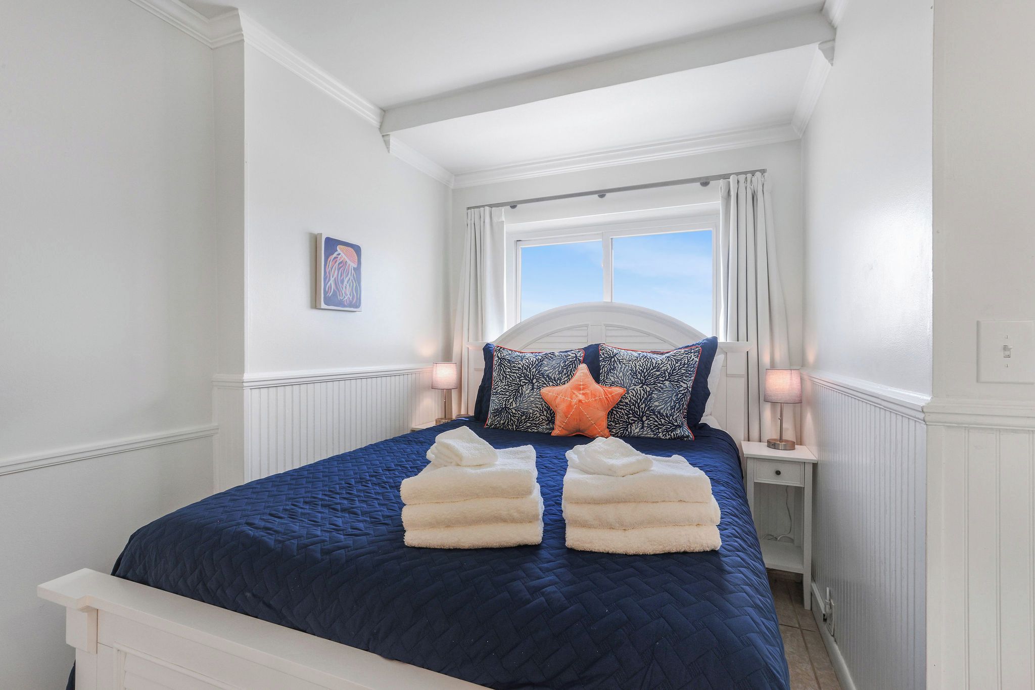 Ein ordentlich bezogenes Bett mit blauer Decke, zwei weißen Handtüchern und dekorativen Kissen in einem Zimmer mit weißen Wänden und einem Fenster.