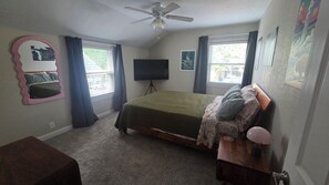 Un dormitorio con cama, ventilador de techo, televisor y ventana con cortinas.