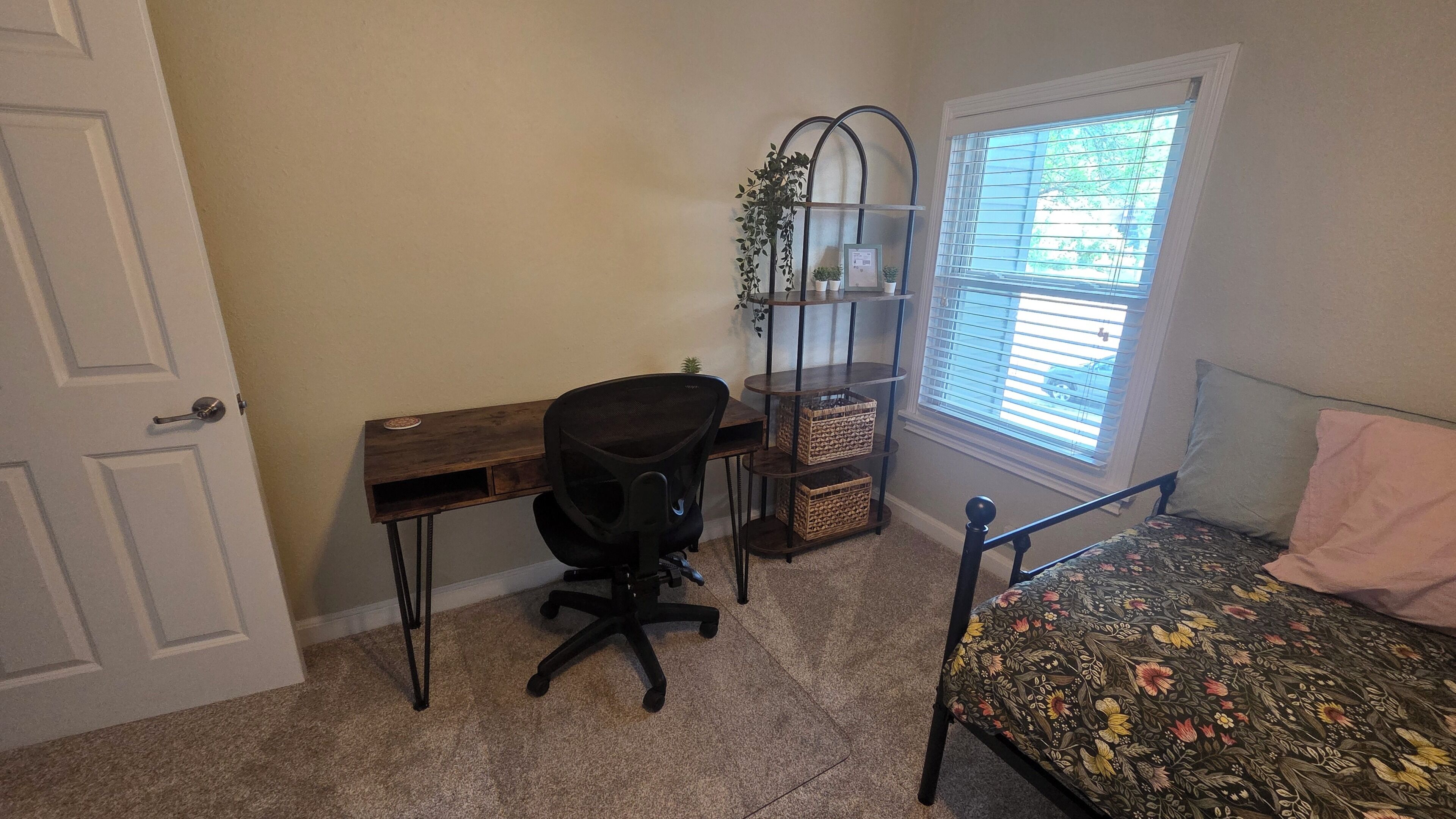 A bedroom with a bed, a desk, a chair, a window with blinds, and a door.