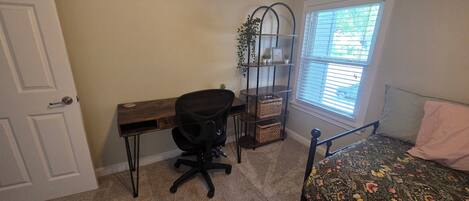 A bedroom with a bed, a desk, a chair, a window with blinds, and a door.