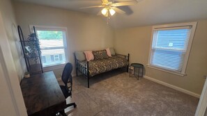 Una habitación con cama, escritorio, silla, ventilador de techo y dos ventanas con persianas.