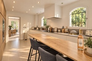 A modern kitchen with a wooden bar, a built-in oven, and a large window with a view of greenery. - Villa Vany - Provence & View (Éguilles)