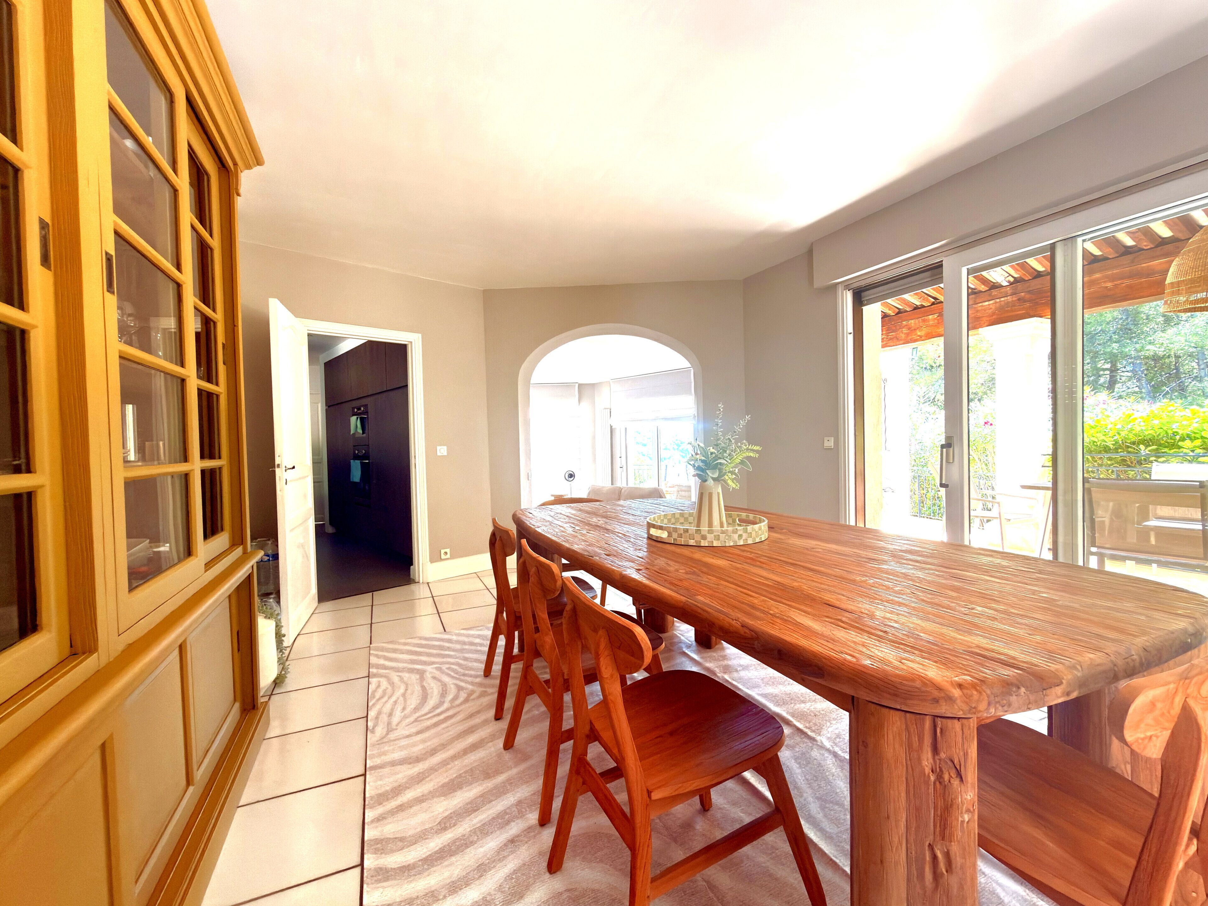 A dining area with a wooden table and chairs, a view of the outdoors through a glass door, and a kitchen visible in the background.